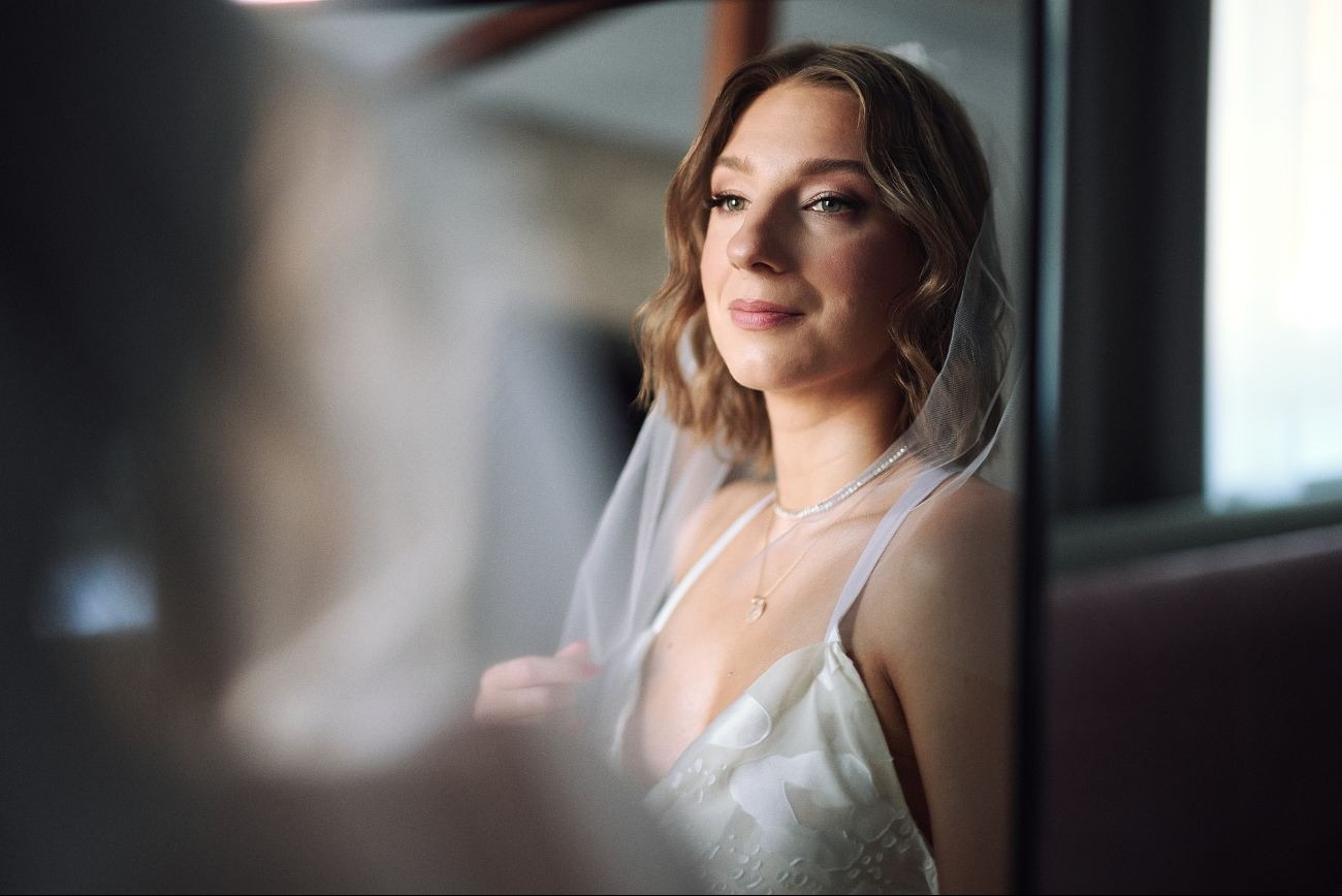 Portret panny młodej w welonie, z delikatnym makijażem i biżuterią, uchwycony w miękkim świetle, z refleksami na sukni ślubnej i subtelnym uśmiechem.