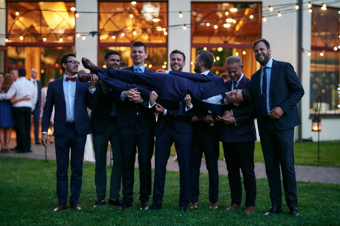 Grupa mężczyzn w garniturach, w tym jeden z czerwoną muszką, podrzuca innego mężczyznę na tle budynku z oknami i dekoracyjnymi lampkami.