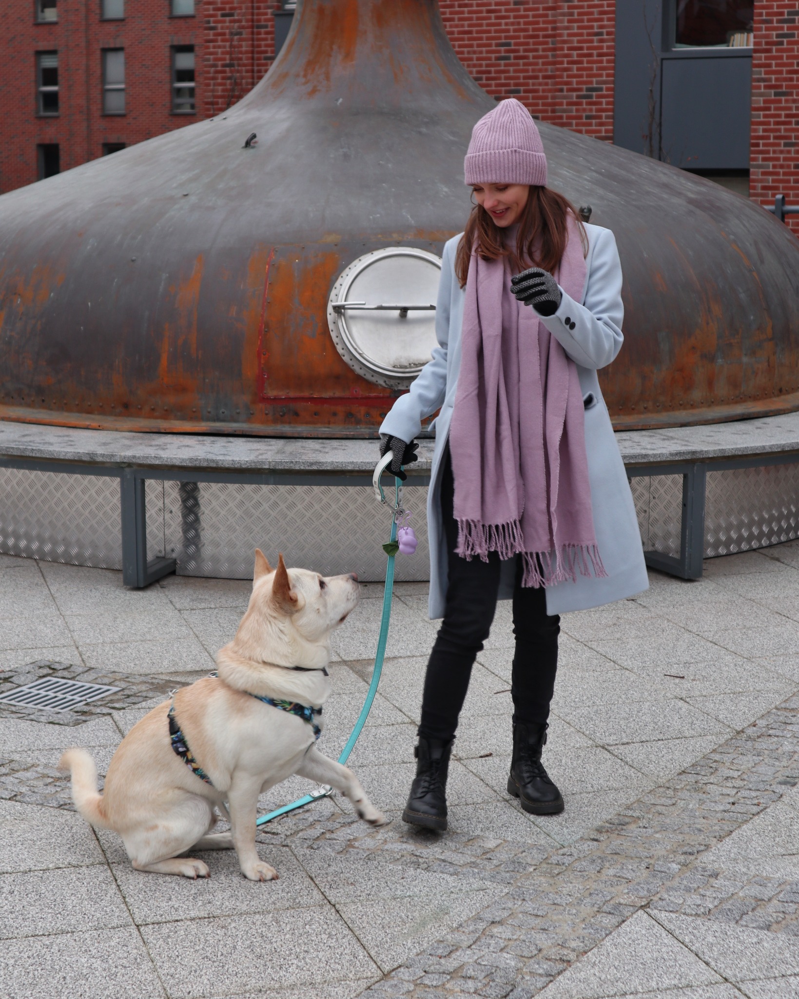 Kobieta w płaszczu i czapce trzyma psa na smyczy. Pies podaje łapę. W tle ceglany budynek i metalowa konstrukcja.