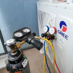 DE Systemy sp. z o.o. - Podłączony zestaw manometrów do klimatyzacji Gree Inverter z butlą z gazem. Widoczne węże w kolorach: żółtym, niebieskim i czerwonym. Sprzęt serwisowy na tle balkonu.