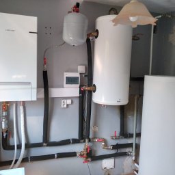 DE Systemy sp. z o.o. - Zestaw urządzeń grzewczych: piec gazowy Samsung, dwa zbiorniki (Galmet, Kospel), naczynie przeponowe, czarne rury w izolacji, szara ściana, lampa.