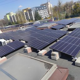 DE Systemy sp. z o.o. - Instalacja paneli słonecznych na płaskim dachu budynku miejskiego, panele podparte na betonowych blokach, widok z góry, słoneczny dzień.