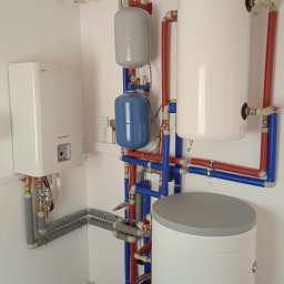 DE Systemy sp. z o.o. - Kotłownia z piecem LG Therma V, zbiornikami Rotenso i instalacją z rur w kolorach czerwonym i niebieskim. Widok na instalację w białym pomieszczeniu.