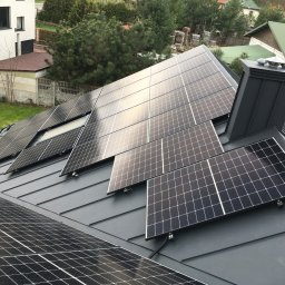 DE Systemy sp. z o.o. - Panele słoneczne na grafitowym dachu domu jednorodzinnego, widok z góry, z kominem i oknem dachowym. W tle zabudowa jednorodzinna i zieleń.