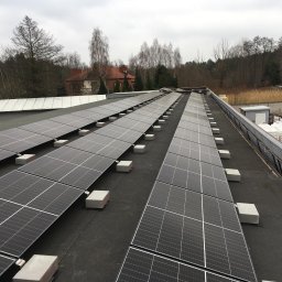 DE Systemy sp. z o.o. - Rząd paneli słonecznych na płaskim dachu budynku, wspartych na betonowych blokach, z widokiem na drzewa i budynki w tle, pochmurne niebo.