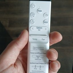 DE Systemy sp. z o.o. - Dłoń trzyma biały pilot Samsung do klimatyzacji, z widocznymi przyciskami sterowania temperaturą, trybem i funkcją Auto Clean na tle drewnianej powierzchni.