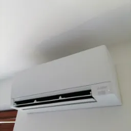 Biała klimatyzacja Mitsubishi Electric zamontowana na jasnej ścianie w pomieszczeniu. Widoczne żaluzje i logo producenta. Minimalistyczny design.