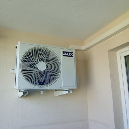 DE Systemy sp. z o.o. - Biała jednostka zewnętrzna klimatyzacji AUX zamontowana na beżowej ścianie budynku, z widocznymi elementami instalacji i fragmentem okna.