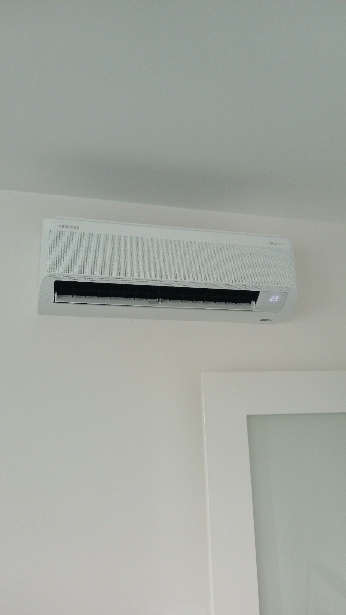 Elegancki, biały klimatyzator Samsung Avant, wyświetlający temperaturę 23°C, zamontowany na ścianie w Warszawie. Widoczny fragment białych drzwi.