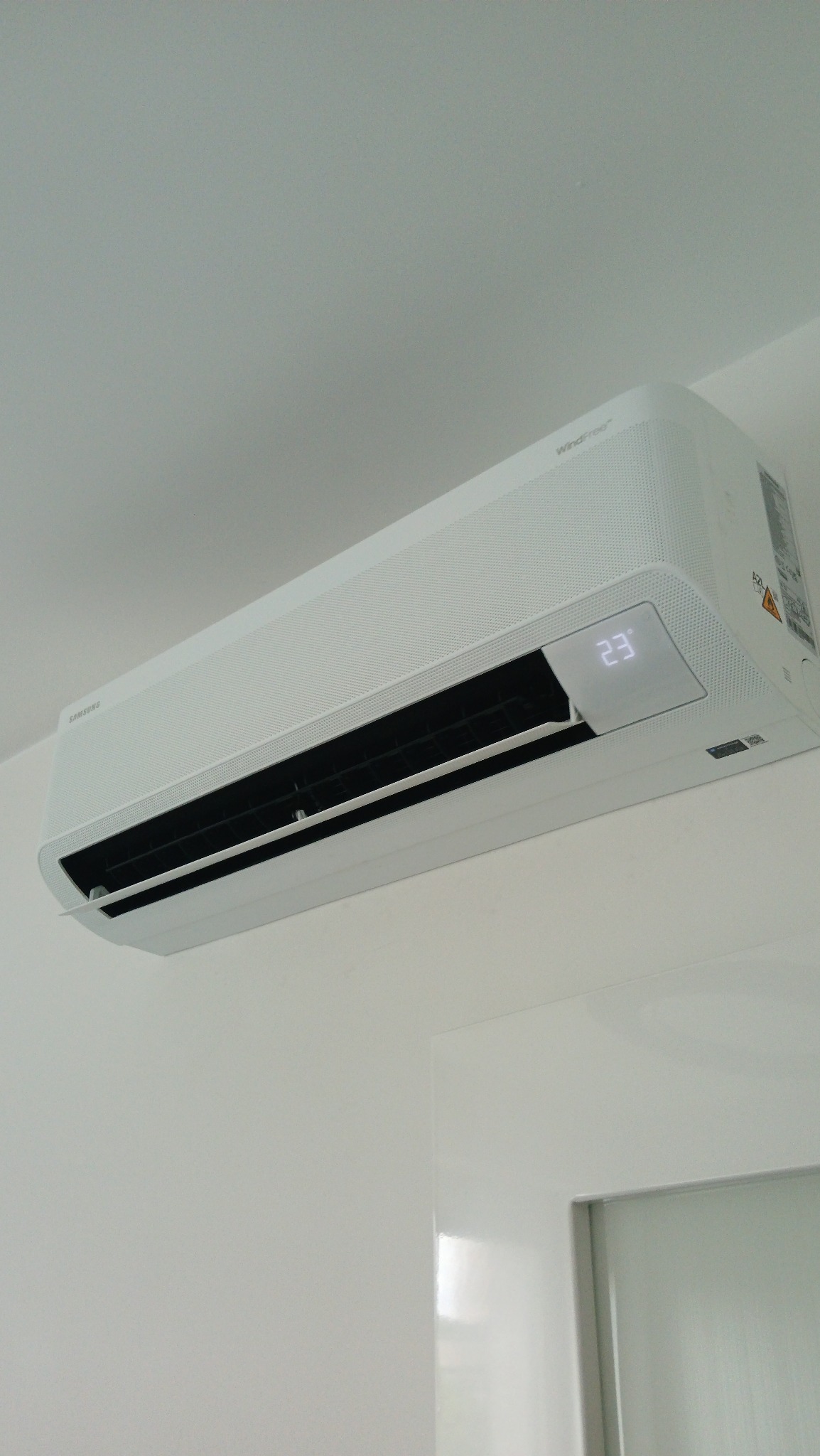 Biały klimatyzator Samsung WindFree zamontowany na ścianie, z wyświetlaczem temperatury 23 stopnie Celsjusza, widok z dołu, fragment drzwi w tle.