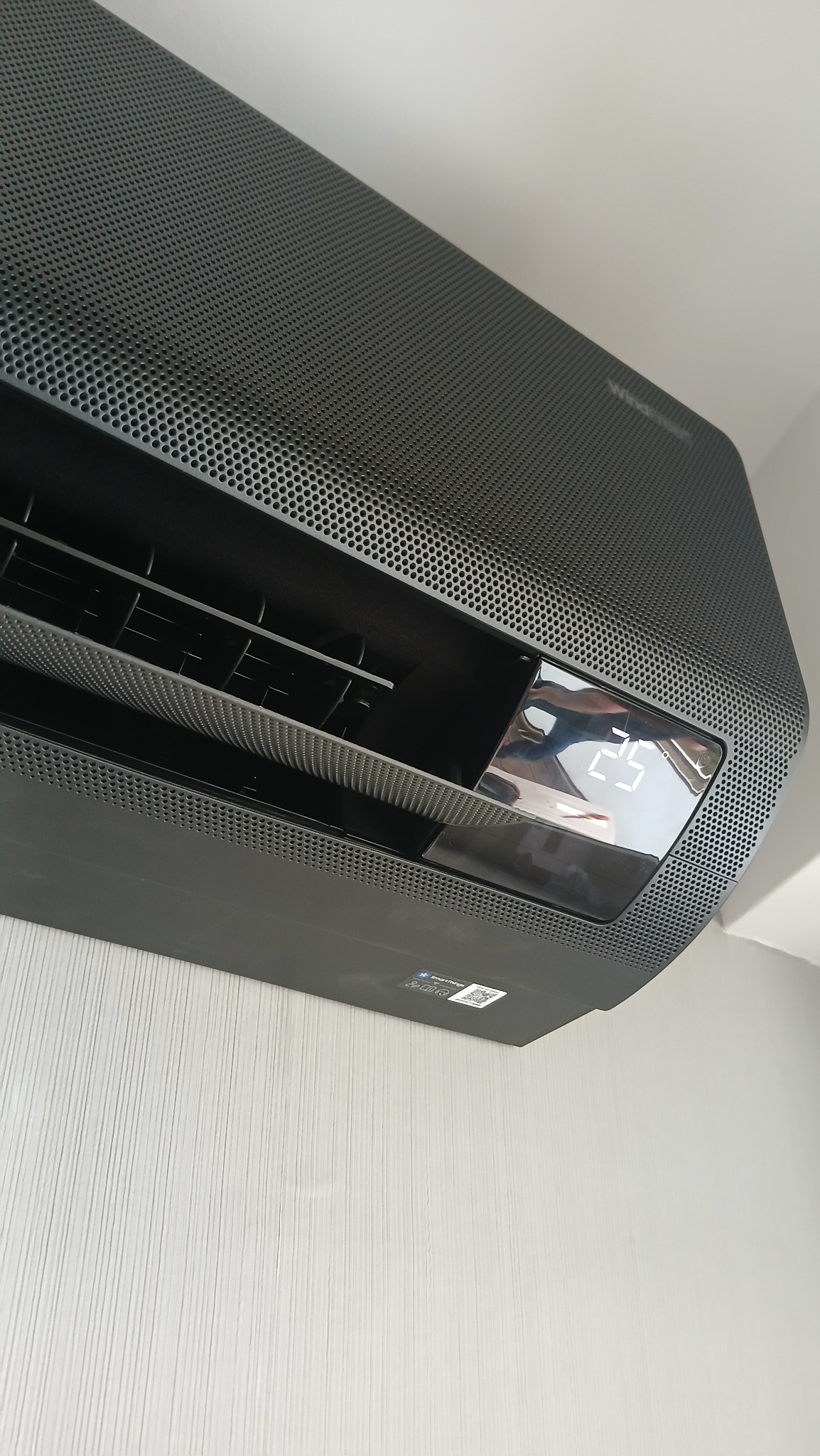 Zamontowany, czarny klimatyzator Samsung WindFree z wyświetlaczem temperatury 25°C na tle ściany z pionowymi paskami. Widoczne perforacje obudowy.