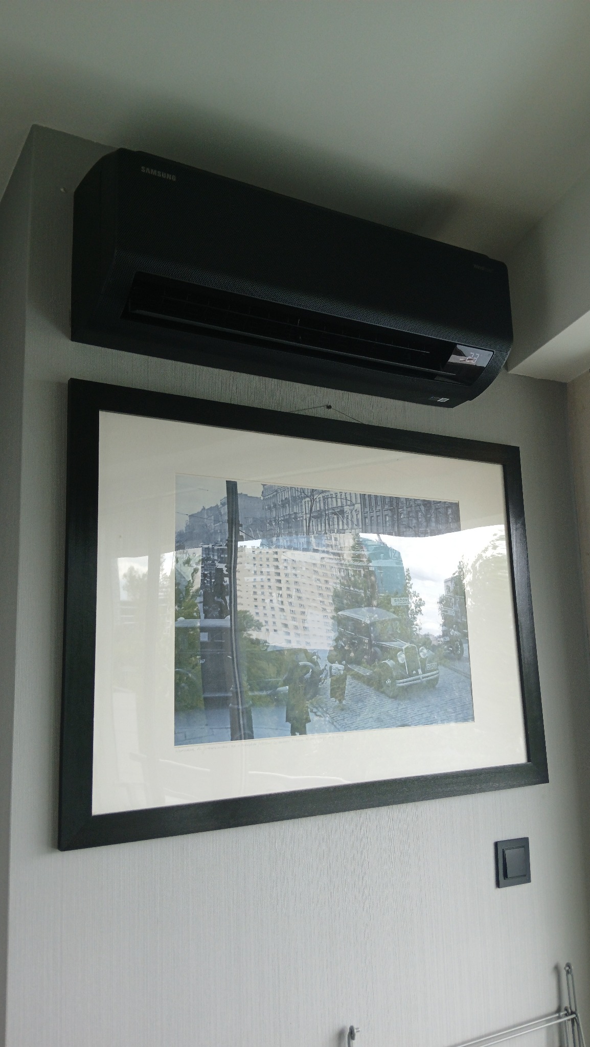 Czarna klimatyzacja Samsung na ścianie nad oprawionym zdjęciem z ulicznym motywem. Nowoczesny design jednostki wewnętrznej w szarej tonacji.