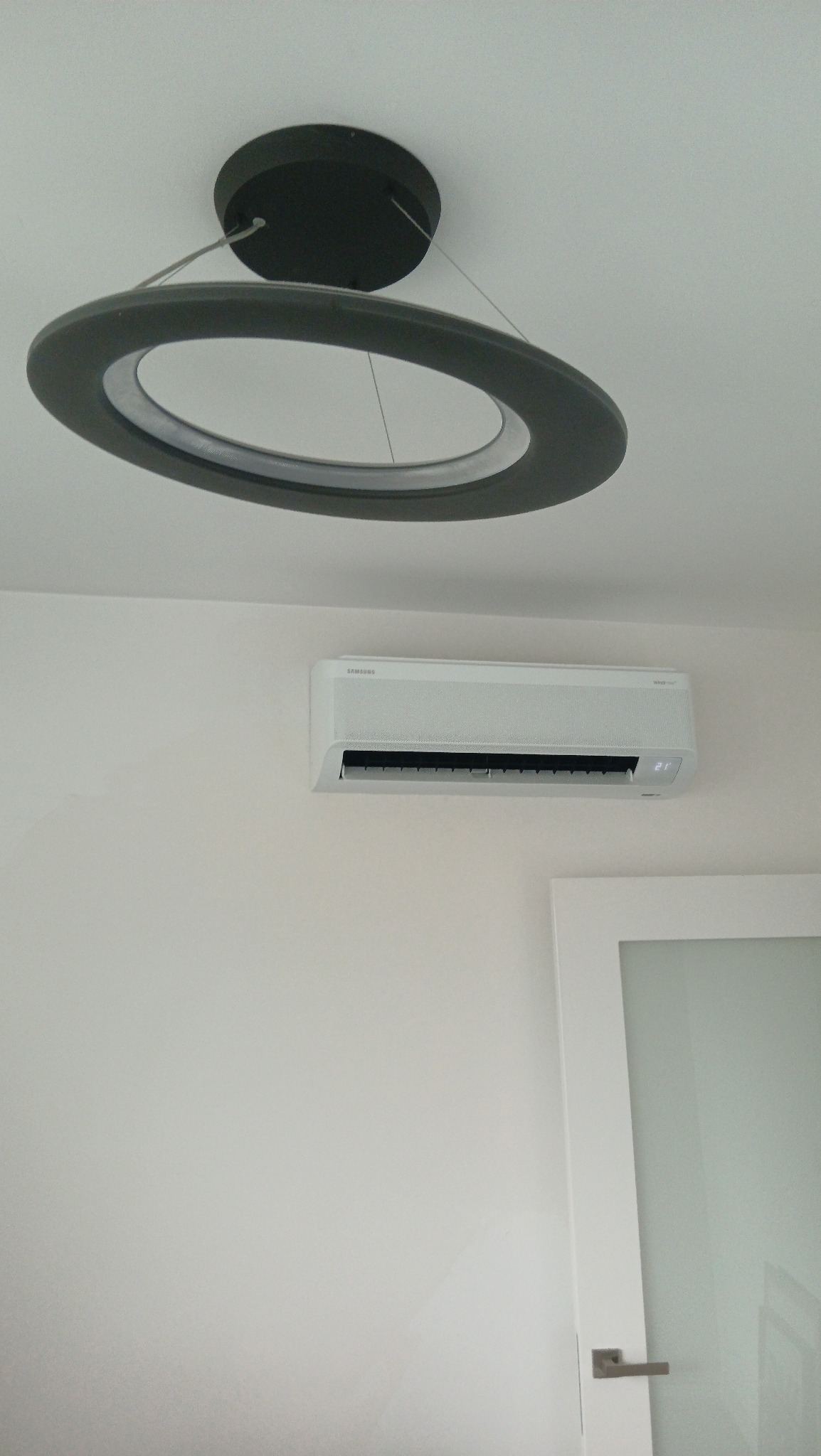 Biały klimatyzator Samsung WindFree zamontowany na ścianie, pod okrągłą lampą sufitową. Widoczny fragment białych drzwi z matową szybą.