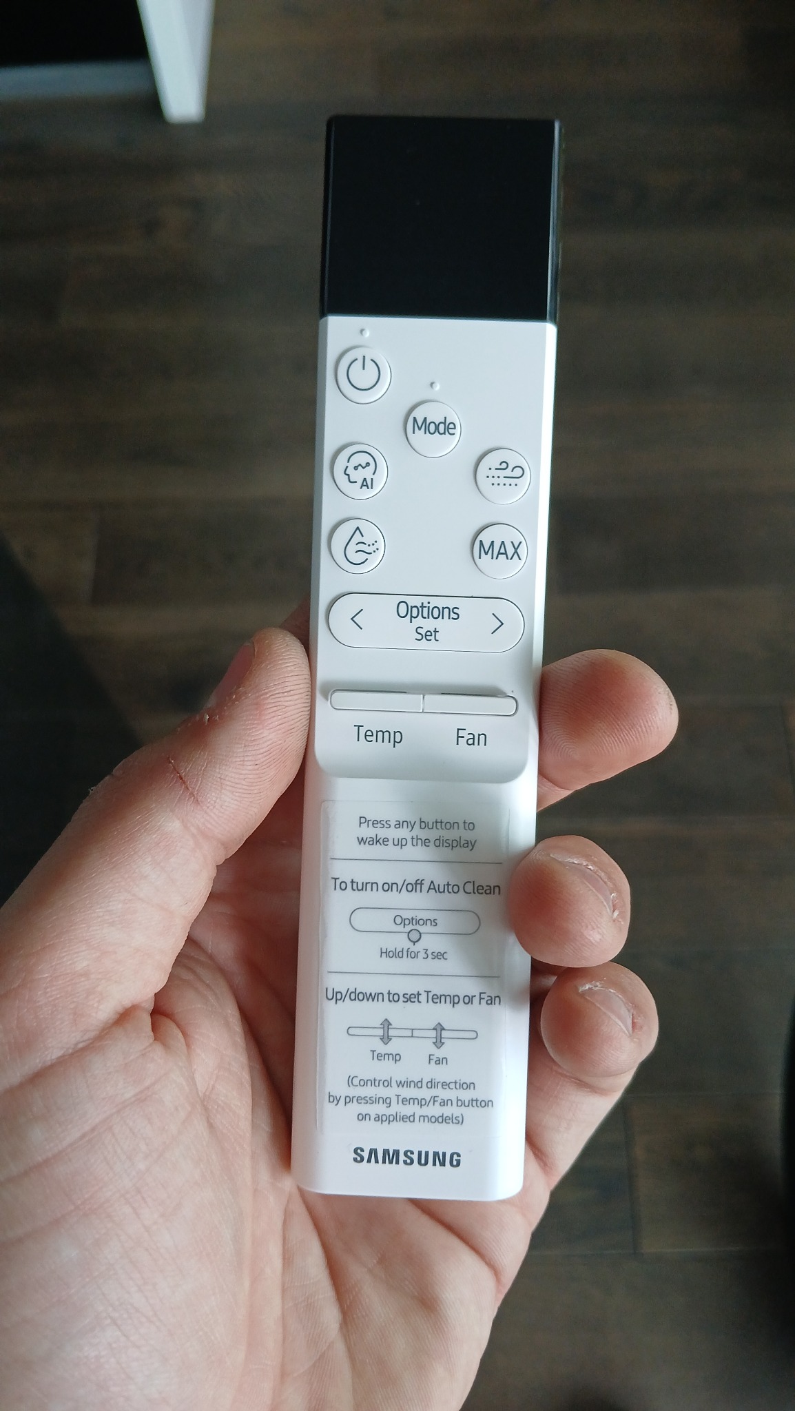 Dłoń trzyma biały pilot Samsung do klimatyzacji, z widocznymi przyciskami sterowania temperaturą, trybem i funkcją Auto Clean na tle drewnianej powierzchni.