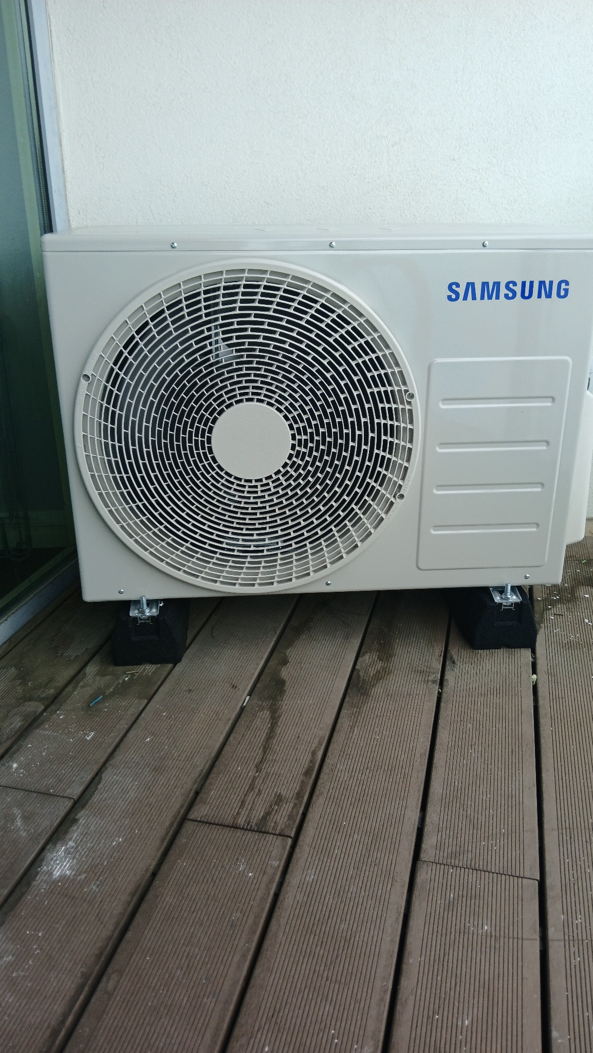 Zewnętrzna jednostka klimatyzacji Samsung na tarasie z desek kompozytowych, zamontowana na czarnych podkładkach antywibracyjnych. Widok z boku.