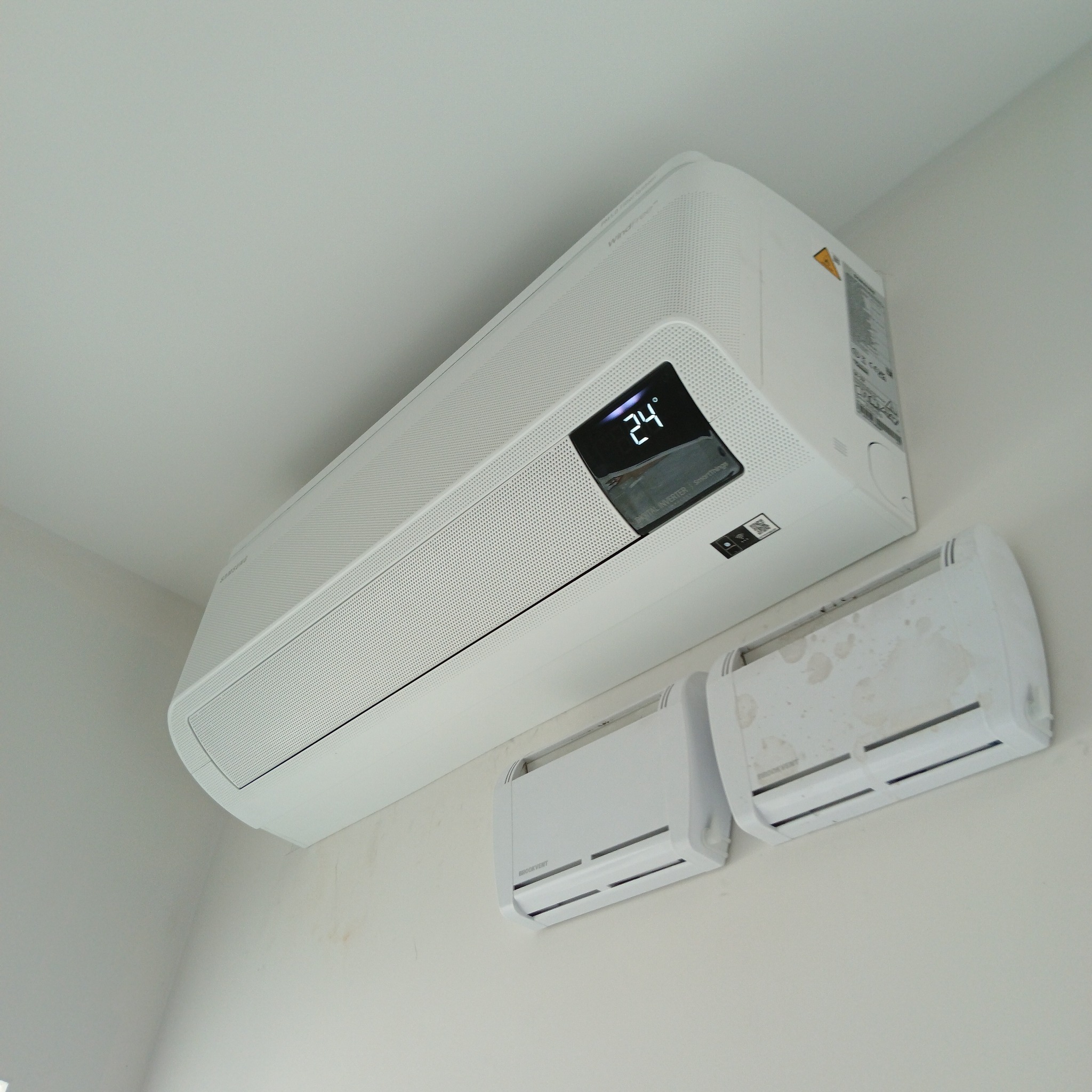 Nowoczesny, biały klimatyzator Samsung z wyświetlaczem 24°C, zamontowany na ścianie obok dwóch mniejszych, prawdopodobnie wentylatorów. Ujęcie z dołu.
