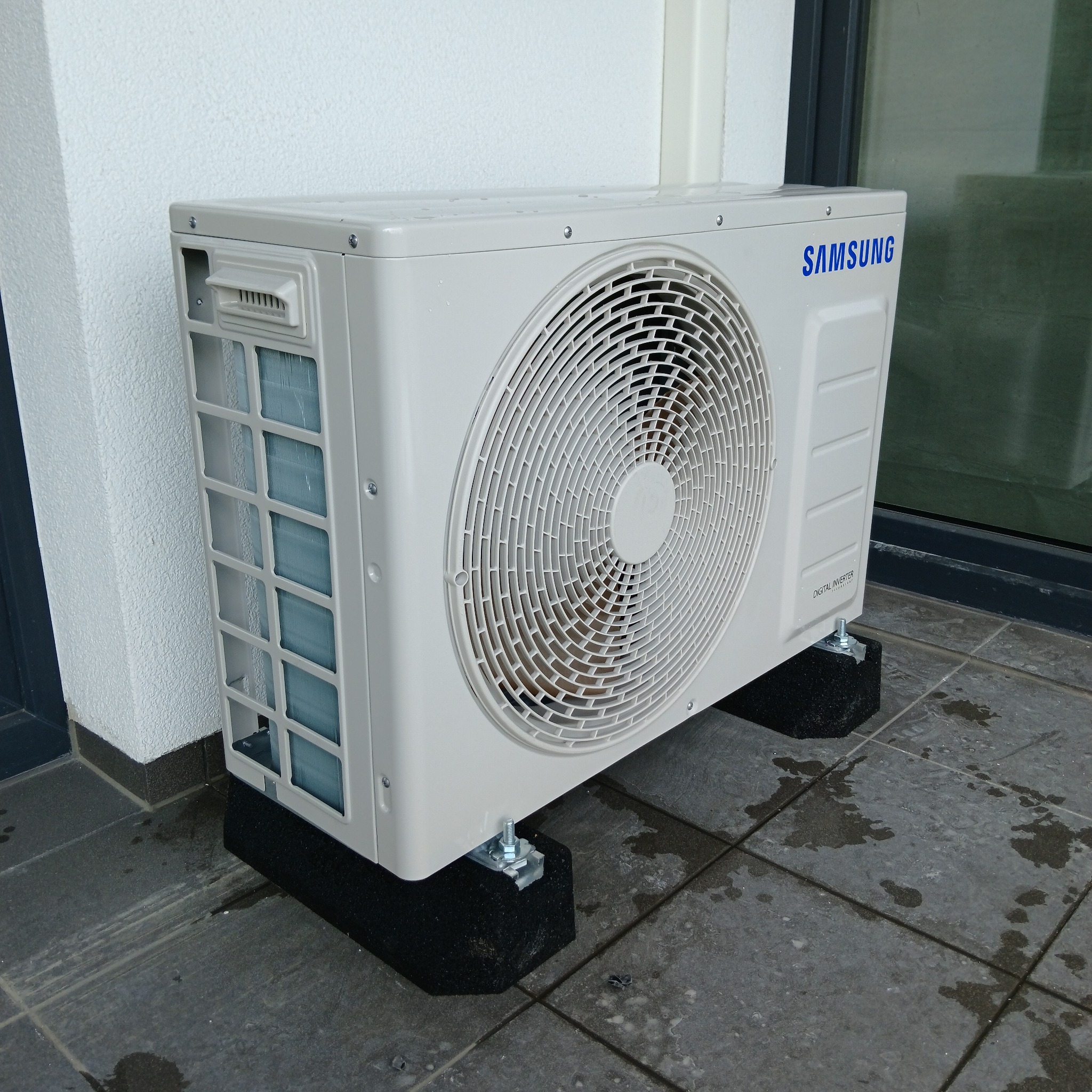 Biała jednostka zewnętrzna klimatyzacji Samsung Digital Inverter, zamontowana na czarnych podkładkach tłumiących drgania, na szarych płytkach tarasu.