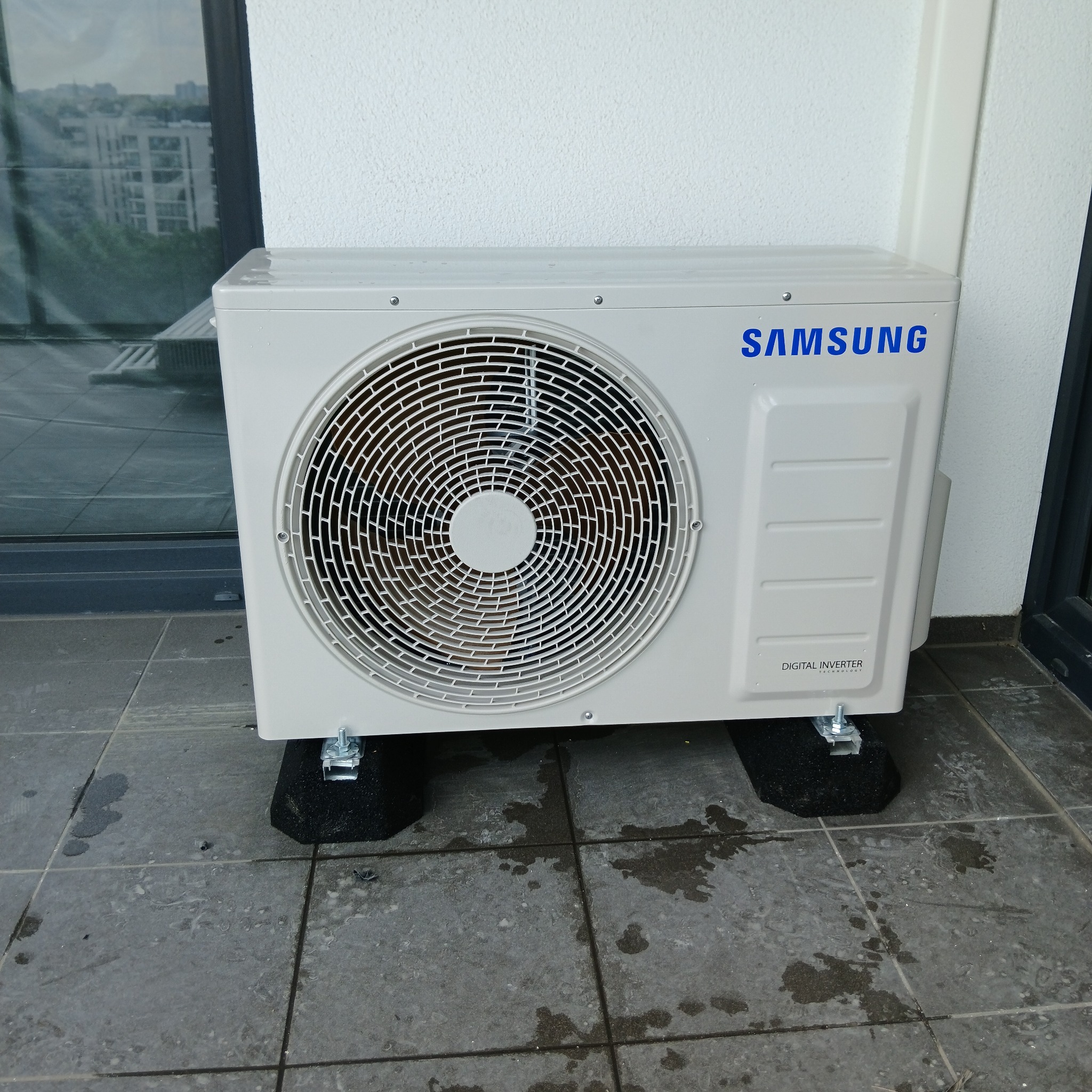 Zewnętrzna jednostka klimatyzacji Samsung Digital Inverter na balkonie, umieszczona na czarnych podkładkach antywibracyjnych. Widoczne ślady wilgoci na podłodze.