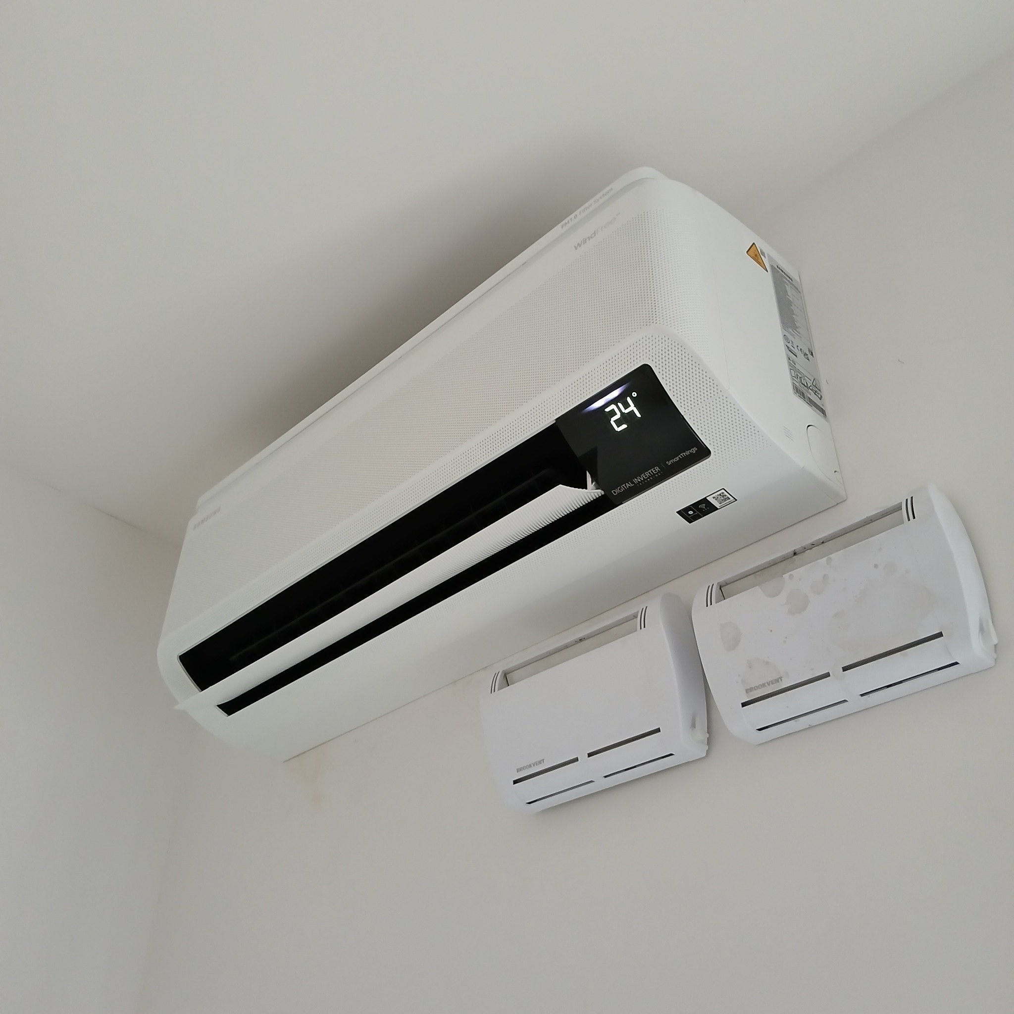 Biała klimatyzacja Samsung WindFree na ścianie, wyświetla temperaturę 24°C. Dodatkowo widoczne dwa białe nawiewniki Brookvent.
