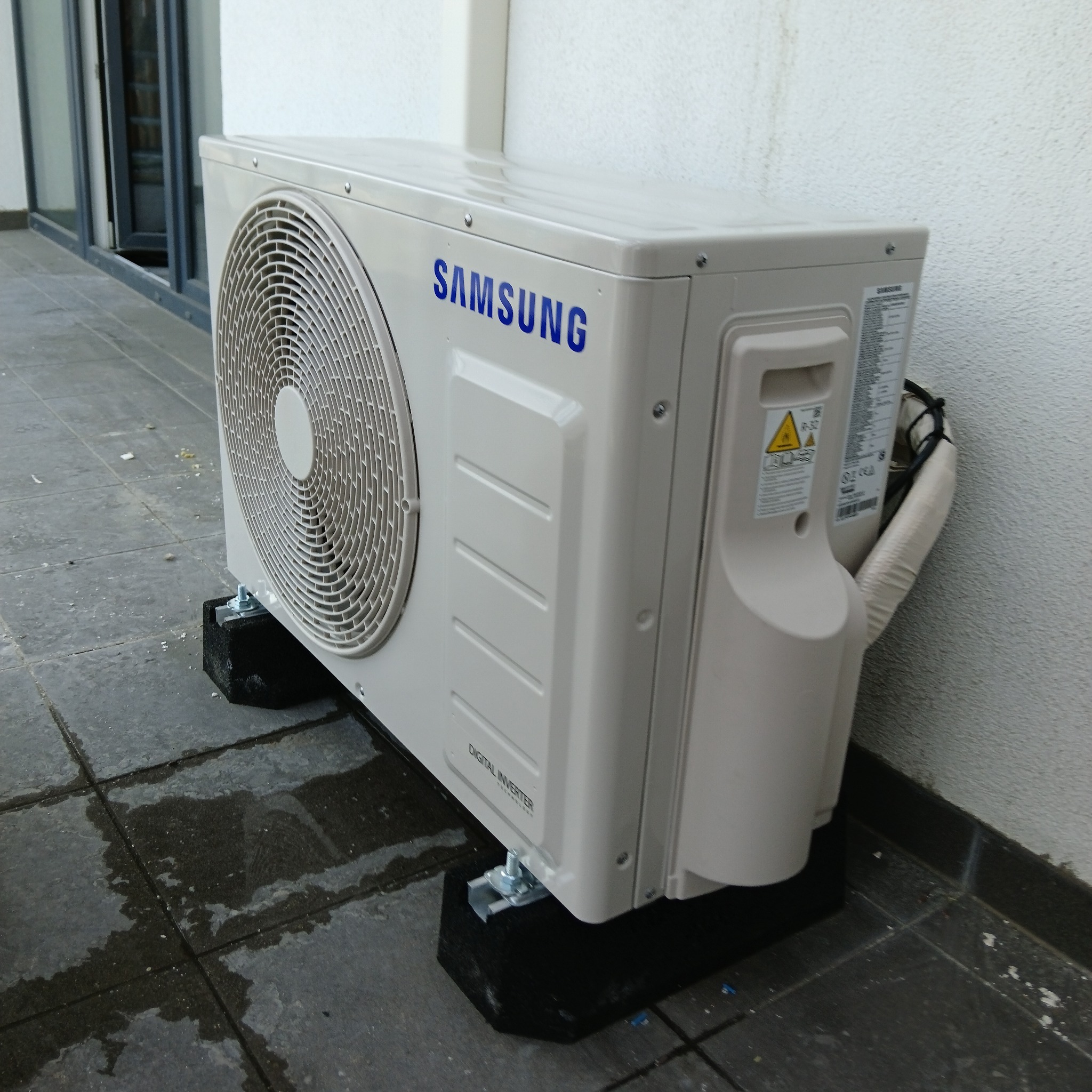 Zewnętrzna jednostka klimatyzacji Samsung na czarnych, gumowych podkładkach, umieszczona na mokrym, szarym tarasie przy białej ścianie budynku.