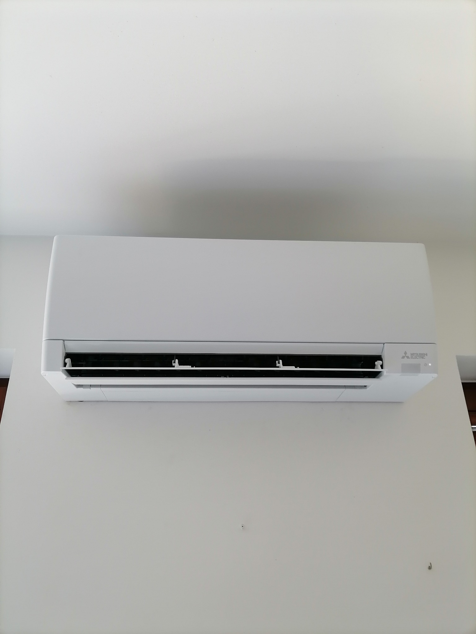 Biały klimatyzator Mitsubishi Electric zamontowany na białej ścianie. Widok z przodu, centralnie. Widoczne detale urządzenia i fragment blatu po prawej stronie.