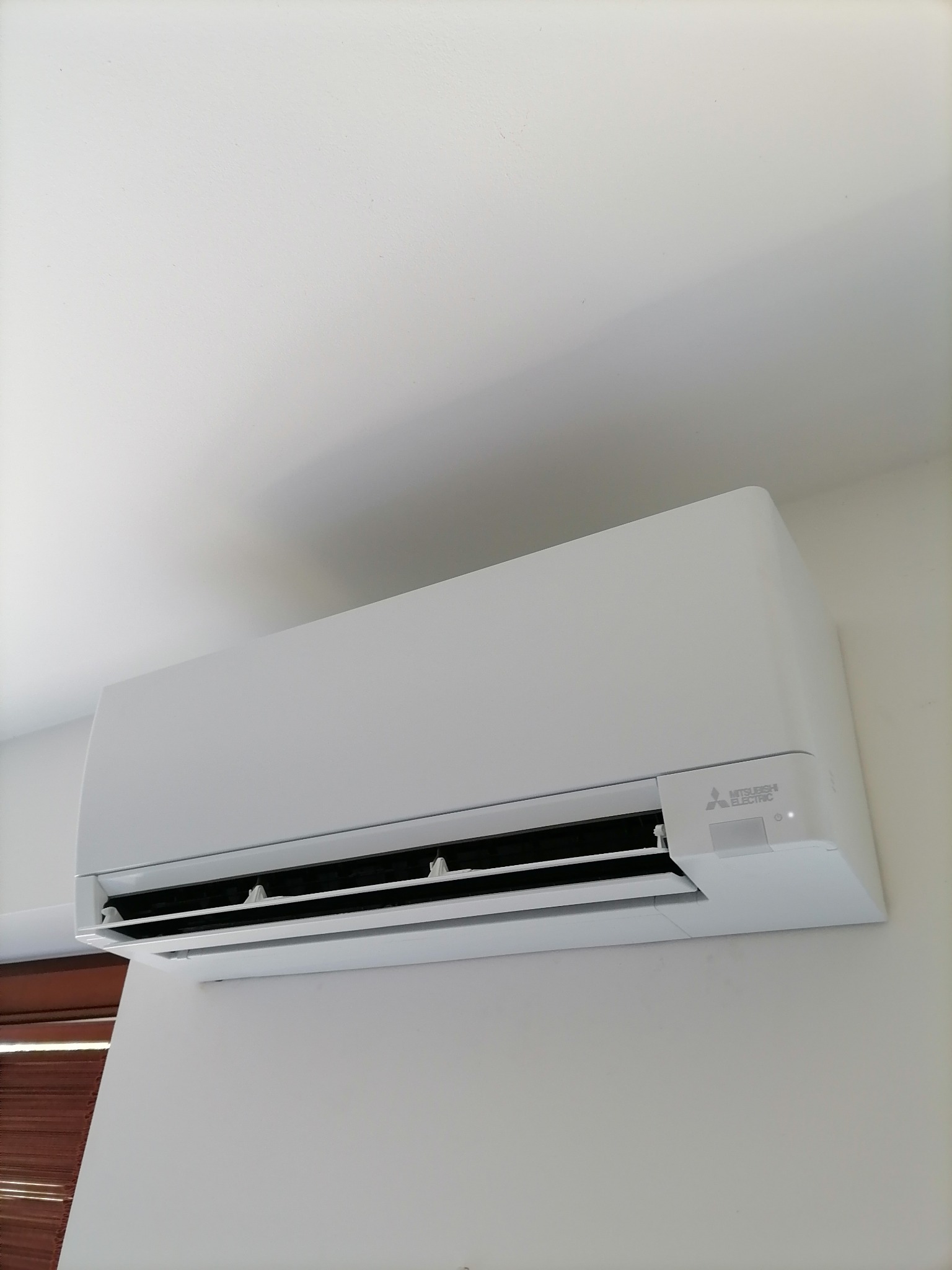 Biała klimatyzacja Mitsubishi Electric zamontowana na jasnej ścianie w pomieszczeniu. Widoczne żaluzje i logo producenta. Minimalistyczny design.