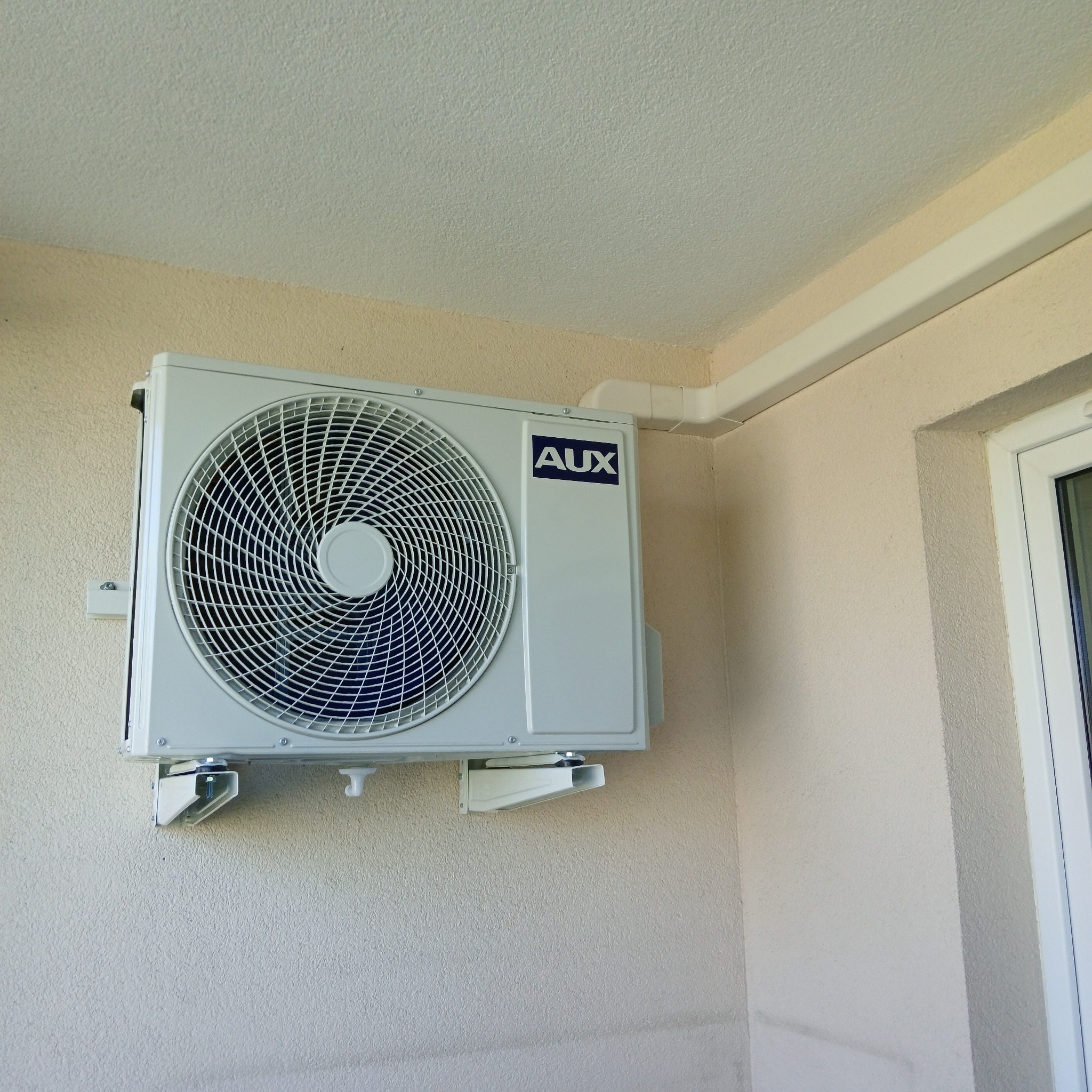 Biała jednostka zewnętrzna klimatyzacji AUX zamontowana na beżowej ścianie budynku, z widocznymi elementami instalacji i fragmentem okna.