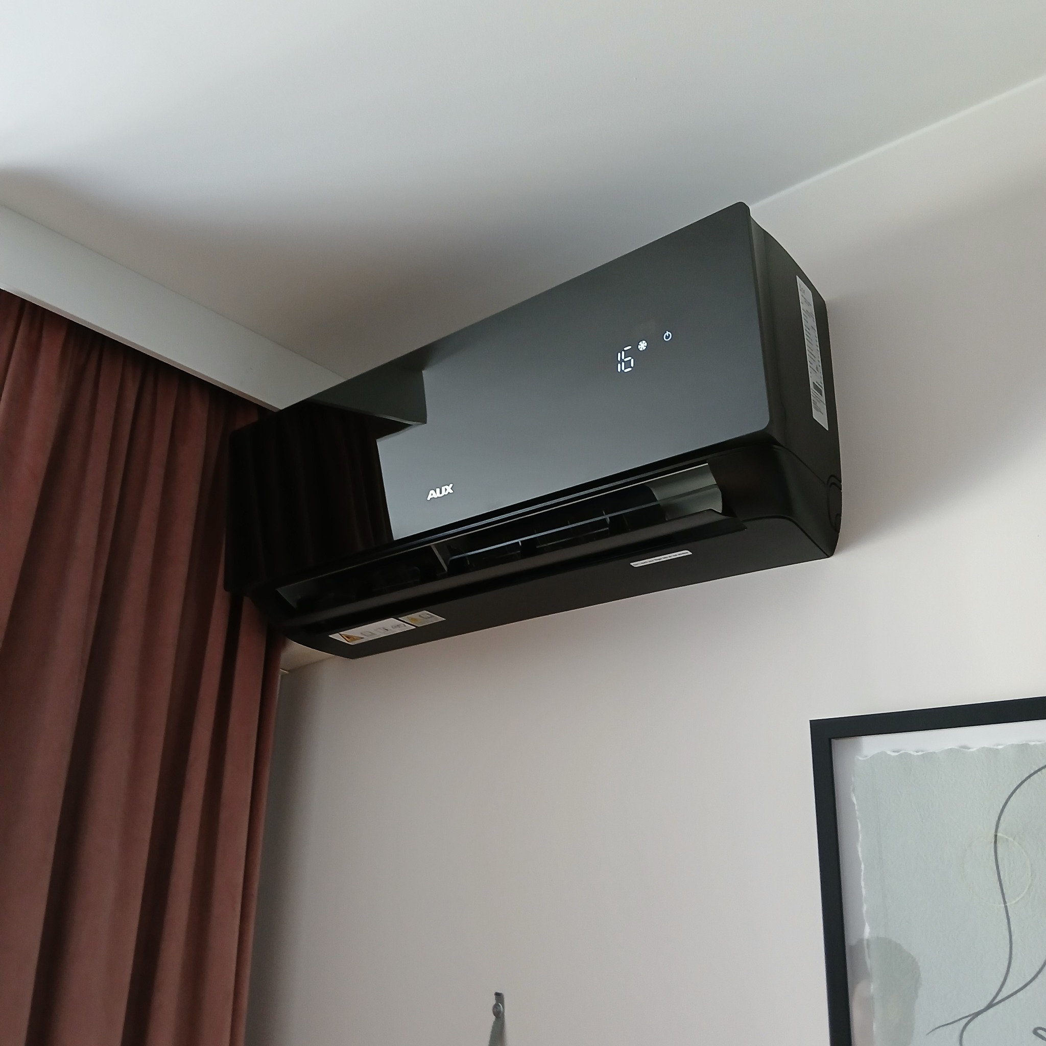 Czarny klimatyzator AUX J-Smart Art zamontowany na ścianie, widoczny wyświetlacz z temperaturą 16 stopni, obok zasłony i obraz w ramie.