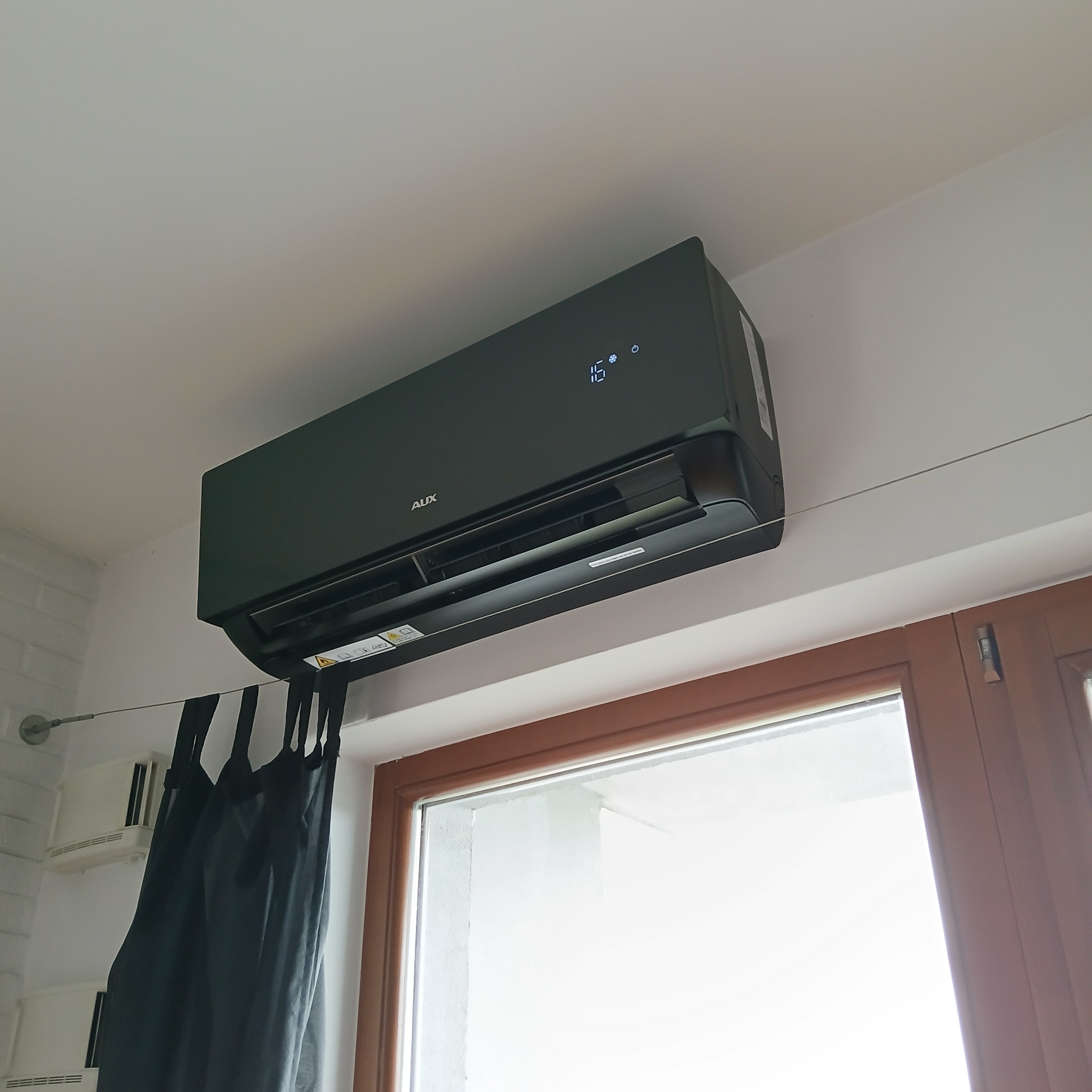 Czarny klimatyzator AUX J-Smart Art zamontowany na ścianie nad oknem, z wyświetlaczem temperatury 16°C. Widoczne zasłony i fragment ściany z cegły.