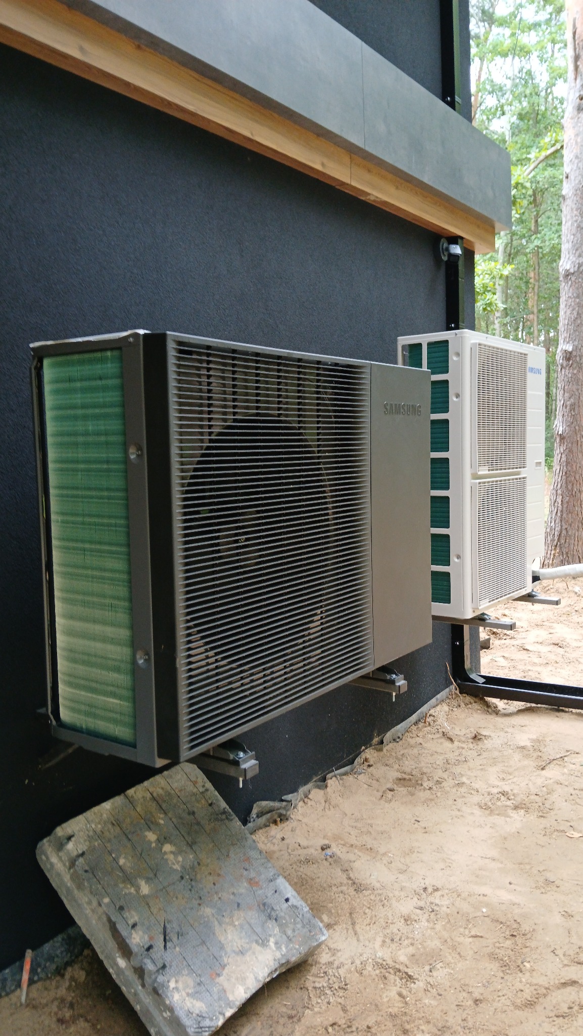 Zewnętrzne jednostki klimatyzacji Samsung, zamontowane na ciemnej elewacji domu. Widoczne elementy montażowe i otoczenie budynku w trakcie prac instalacyjnych.