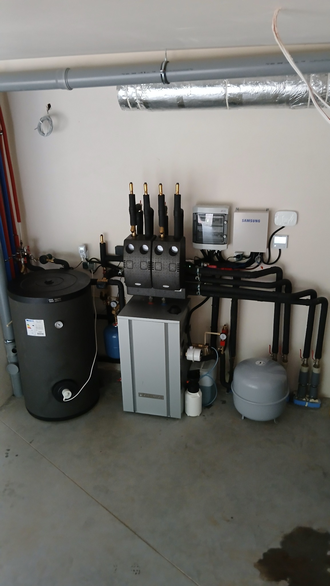 Kompletna instalacja pompy ciepła w kotłowni, z szarym zbiornikiem buforowym i modułem hydraulicznym Wolf. Widoczne rury, zawory i elementy sterujące.