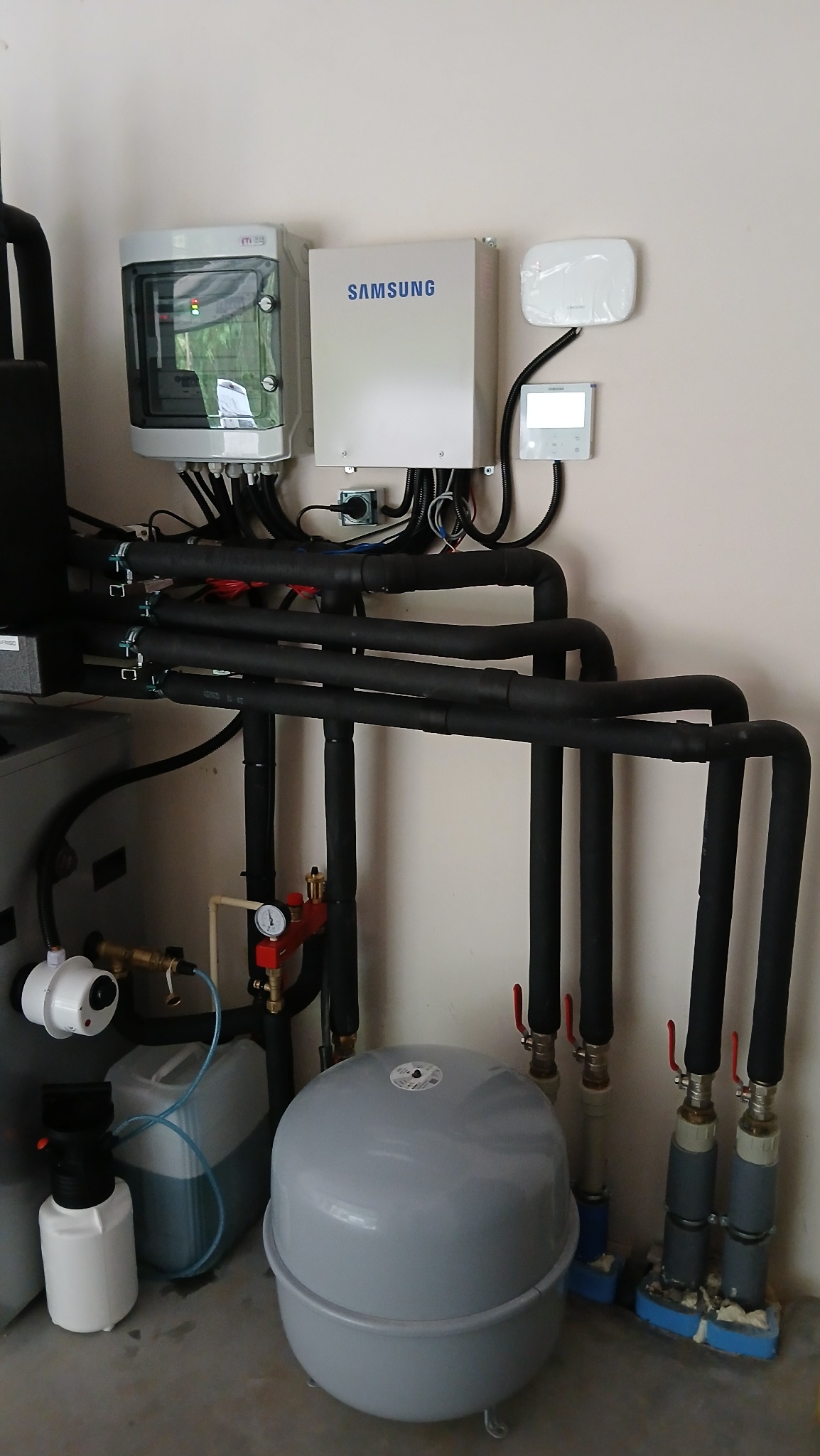 Złożona instalacja grzewcza z czarnymi rurami, zbiornikiem wyrównawczym i elektroniką Samsung na ścianie. Elementy systemu w pomieszczeniu technicznym.