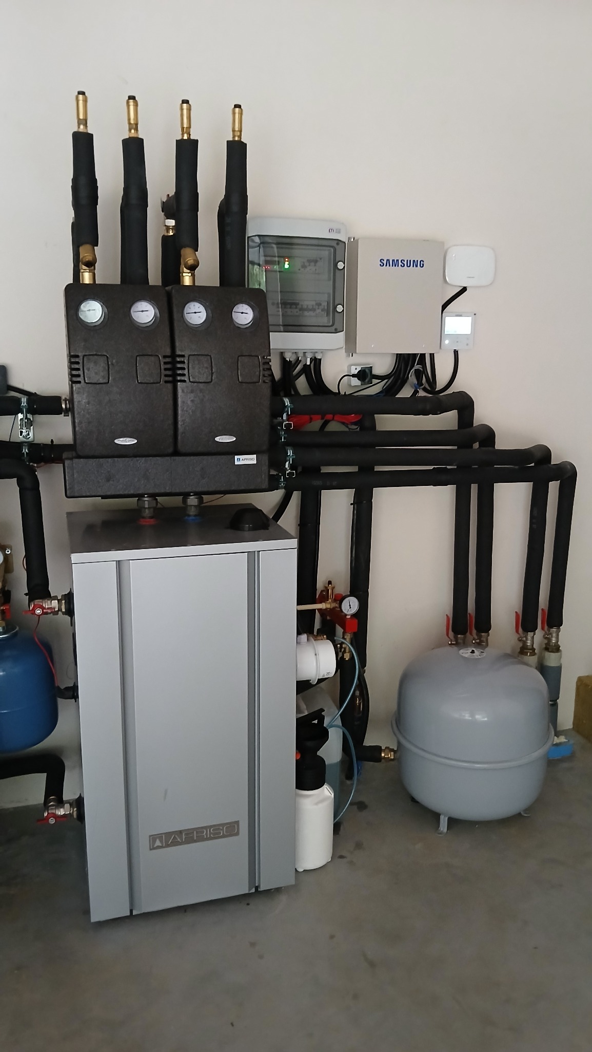 Kompletna instalacja grzewcza z piecem AFRISO, czarnymi rurami, zbiornikiem wyrównawczym i osprzętem, zamontowana przy białej ścianie w pomieszczeniu.