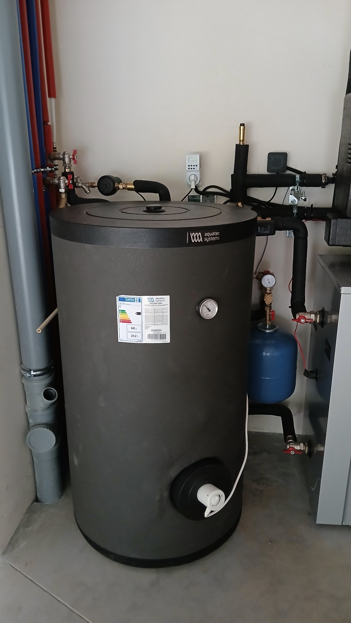 Szary, cylindryczny zbiornik Aquatec Systems z manometrem i etykietą energetyczną, stojący w rogu pomieszczenia z rurami instalacyjnymi.
