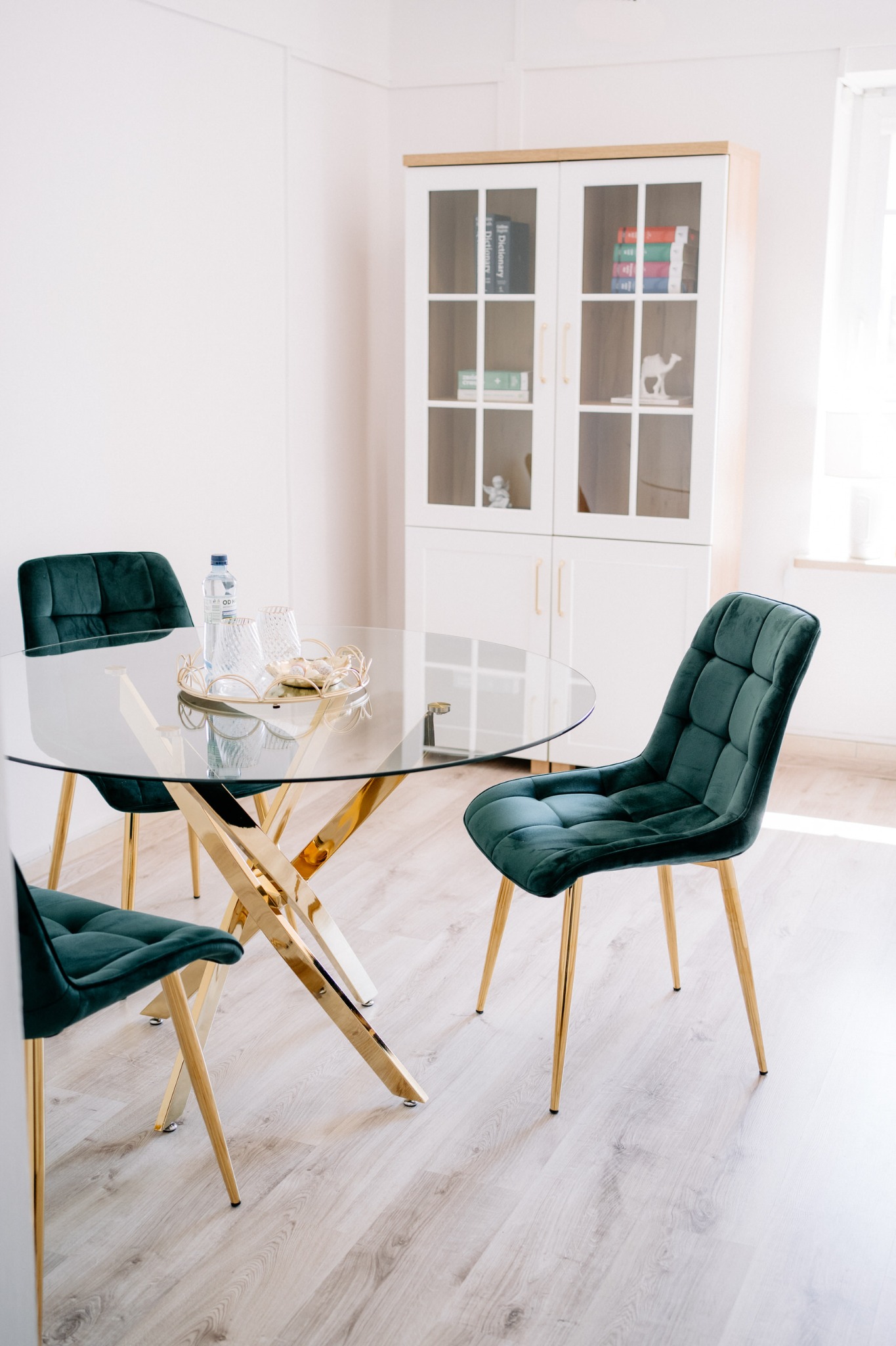 Szklany stół na złotej podstawie otoczony trzema krzesłami z zielonym obiciem. W tle biała szafka z książkami i dekoracjami. Jasne wnętrze.