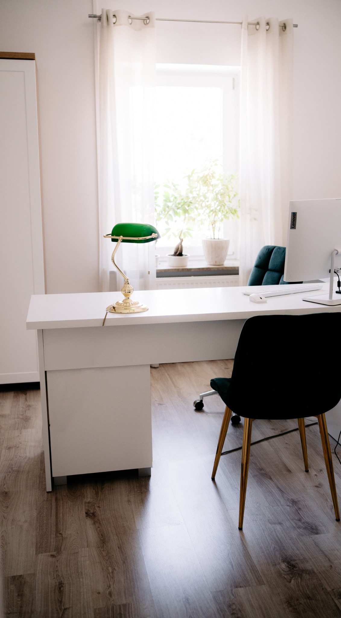 Jasne biuro adwokackie: białe biurko z zieloną lampą bankierską, komputer i krzesło na tle okna z rośliną. Minimalistyczny design.