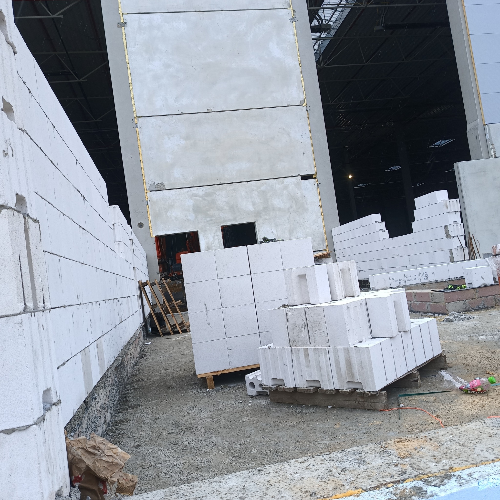 Wznoszenie ścian z białych bloczków betonu komórkowego Ytong na budowie. Widoczne palety z materiałem i fragmenty konstrukcji budynku w tle.