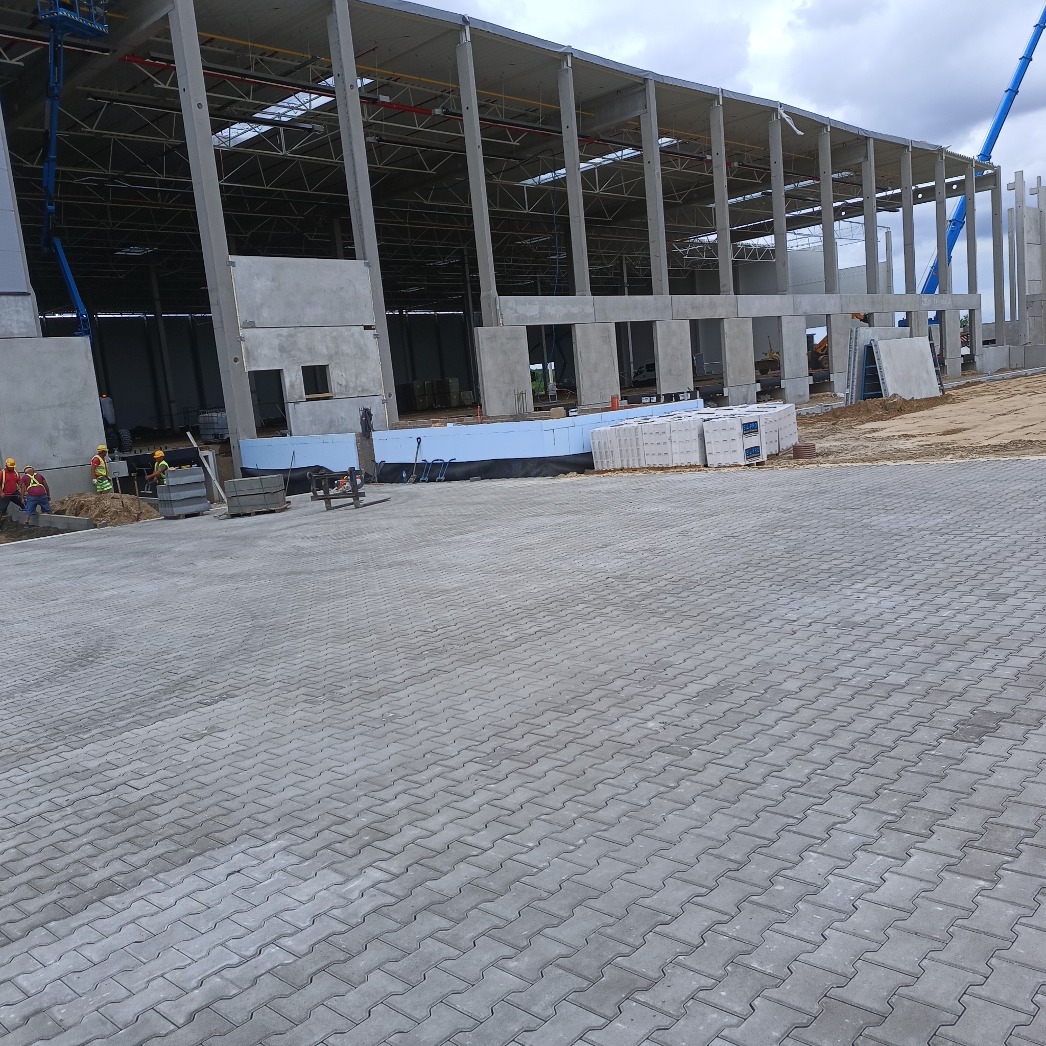 Konstrukcja hali z żelbetu w trakcie budowy. Widoczne szkieletowe elementy, izolacja, palety z materiałami i pracownicy w odblaskowych ubraniach.