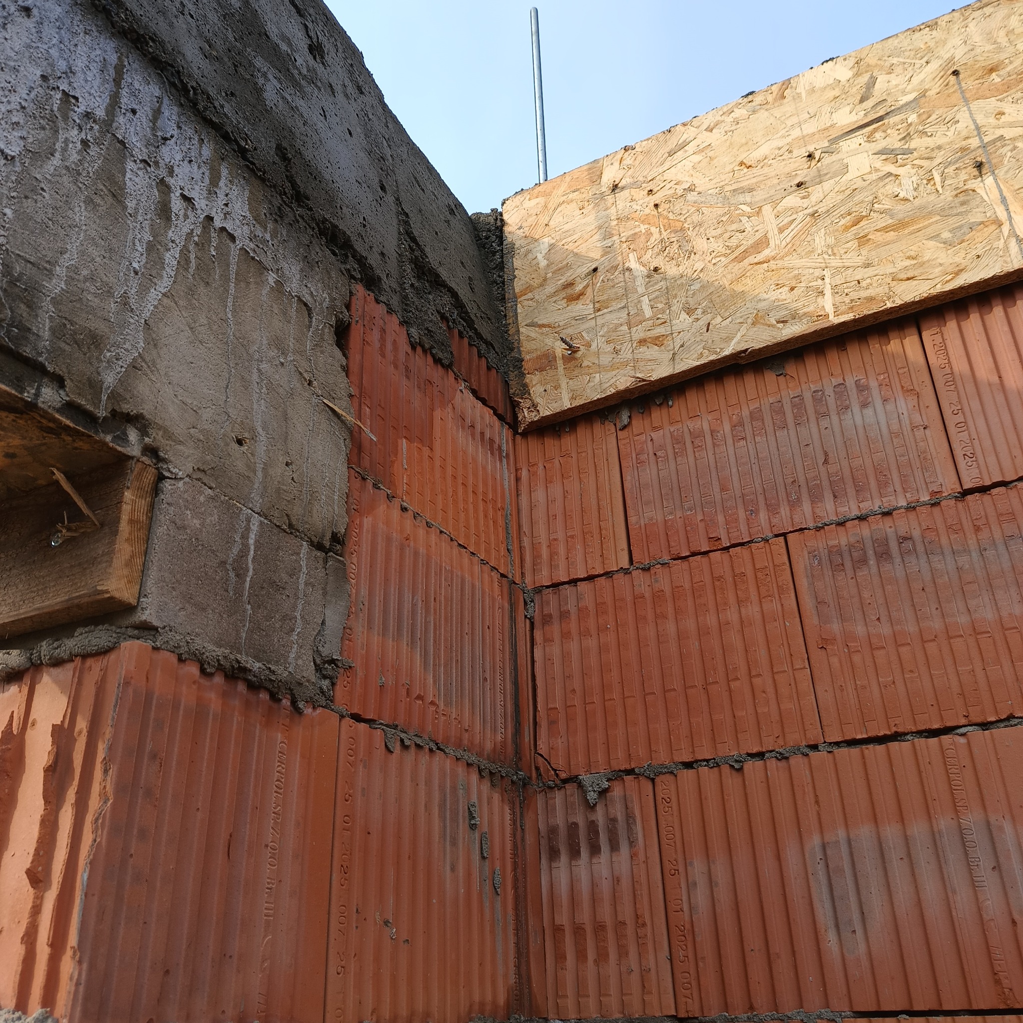 Wznoszenie muru z czerwonej ceramiki poryzowanej, widoczny fragment betonowego szalunku i deski szalunkowej. Etap budowy domu, detal narożnika.