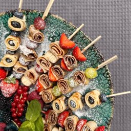 Szef Kuchni Poleca - Dekoracyjne szaszłyki z naleśników, owoców i cukru pudru na talerzu z zielonymi zdobieniami. Widoczne maliny, truskawki, borówki i listki mięty.