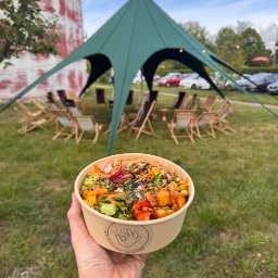 Szef Kuchni Poleca - Ręka trzyma miskę z poke bowl na tle zielonego namiotu i leżaków na trawie. Widoczny budynek w tle. Event na świeżym powietrzu z jedzeniem.