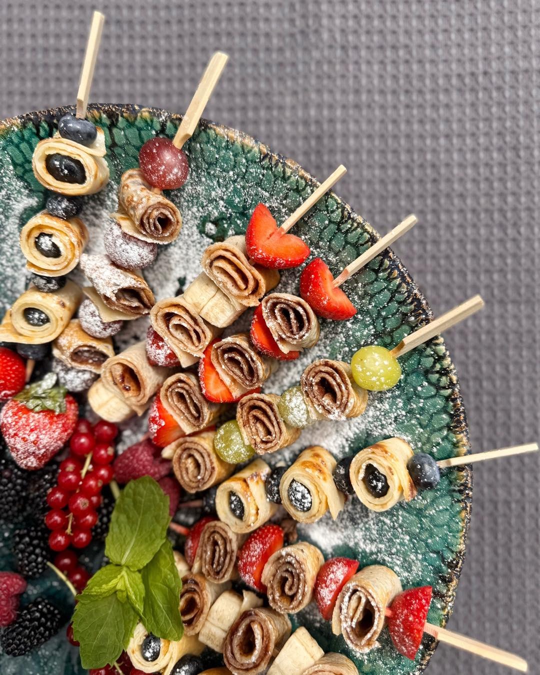 Dekoracyjne szaszłyki z naleśników, owoców i cukru pudru na talerzu z zielonymi zdobieniami. Widoczne maliny, truskawki, borówki i listki mięty.