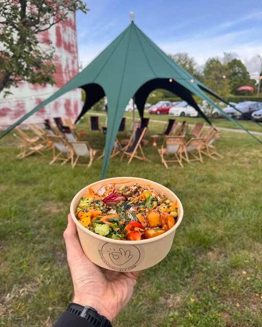 Ręka trzyma miskę z poke bowl na tle zielonego namiotu i leżaków na trawie. Widoczny budynek w tle. Event na świeżym powietrzu z jedzeniem.