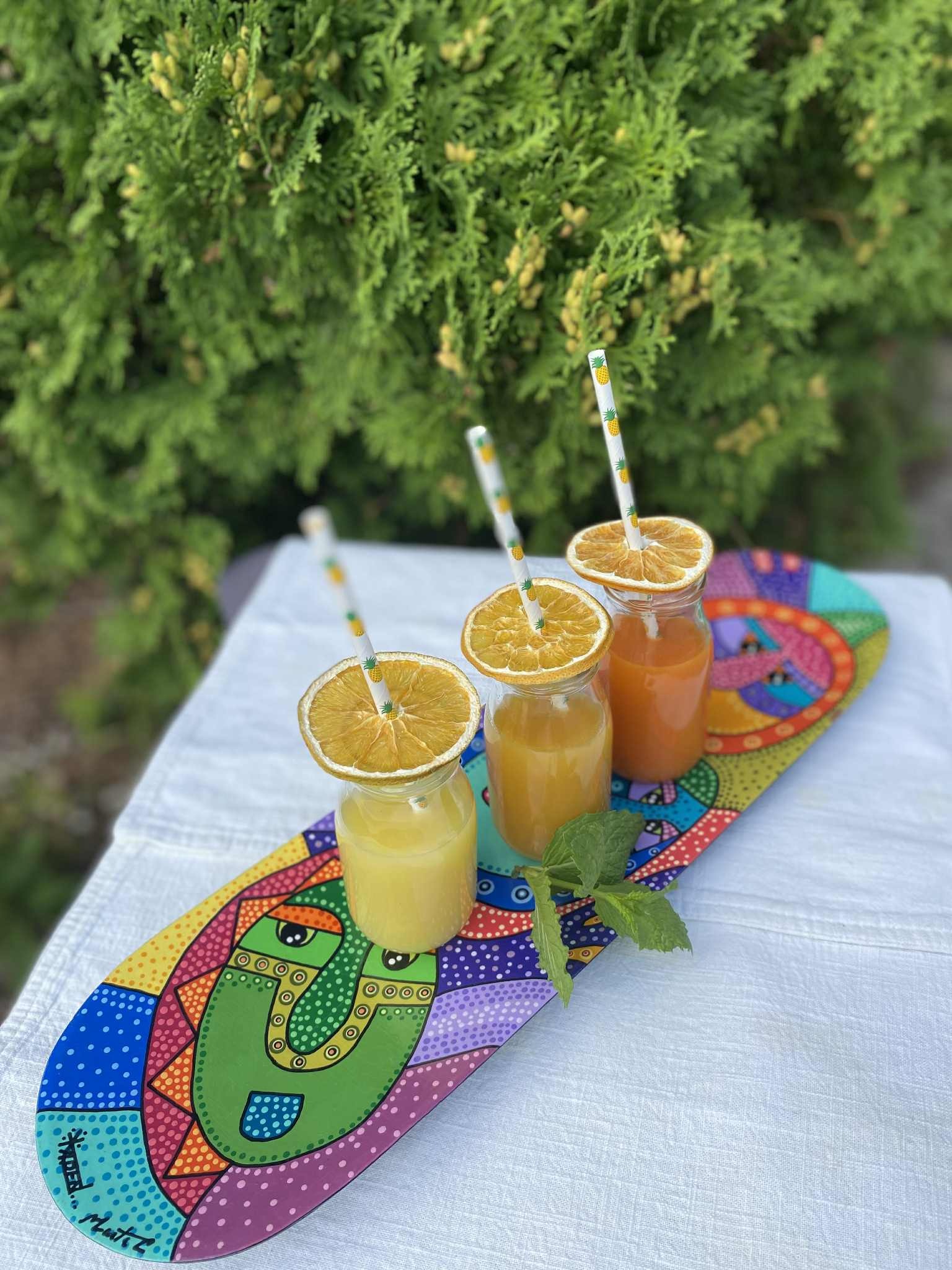 Trzy szklane słoiczki z kolorowymi sokami, udekorowane suszoną pomarańczą i słomkami z ananasami, stoją na ręcznie malowanej desce z motywem twarzy na białym obrusie.