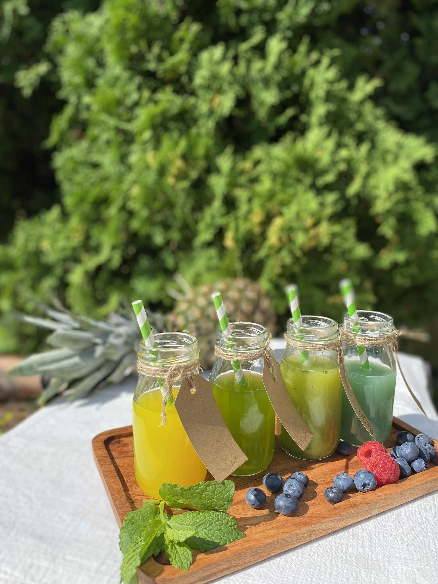 Cztery szklane butelki z kolorowymi sokami, ozdobione sznurkiem i etykietami, na drewnianej tacy z borówkami i malinami. W tle ananas i zielona roślinność.
