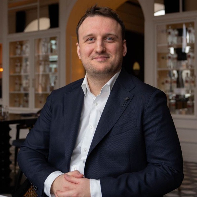 Mężczyzna w ciemnym garniturze i białej koszuli, z delikatnym uśmiechem, siedzi na tle przeszklonej witryny. Dłonie złożone przed sobą, subtelny detal w klapie marynarki.