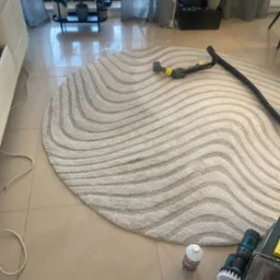 Okrągły dywan w fale czyszczony odkurzaczem piorącym. Na podłodze butelka płynu i szczotka do czyszczenia. Widok z góry, oświetlenie dzienne.