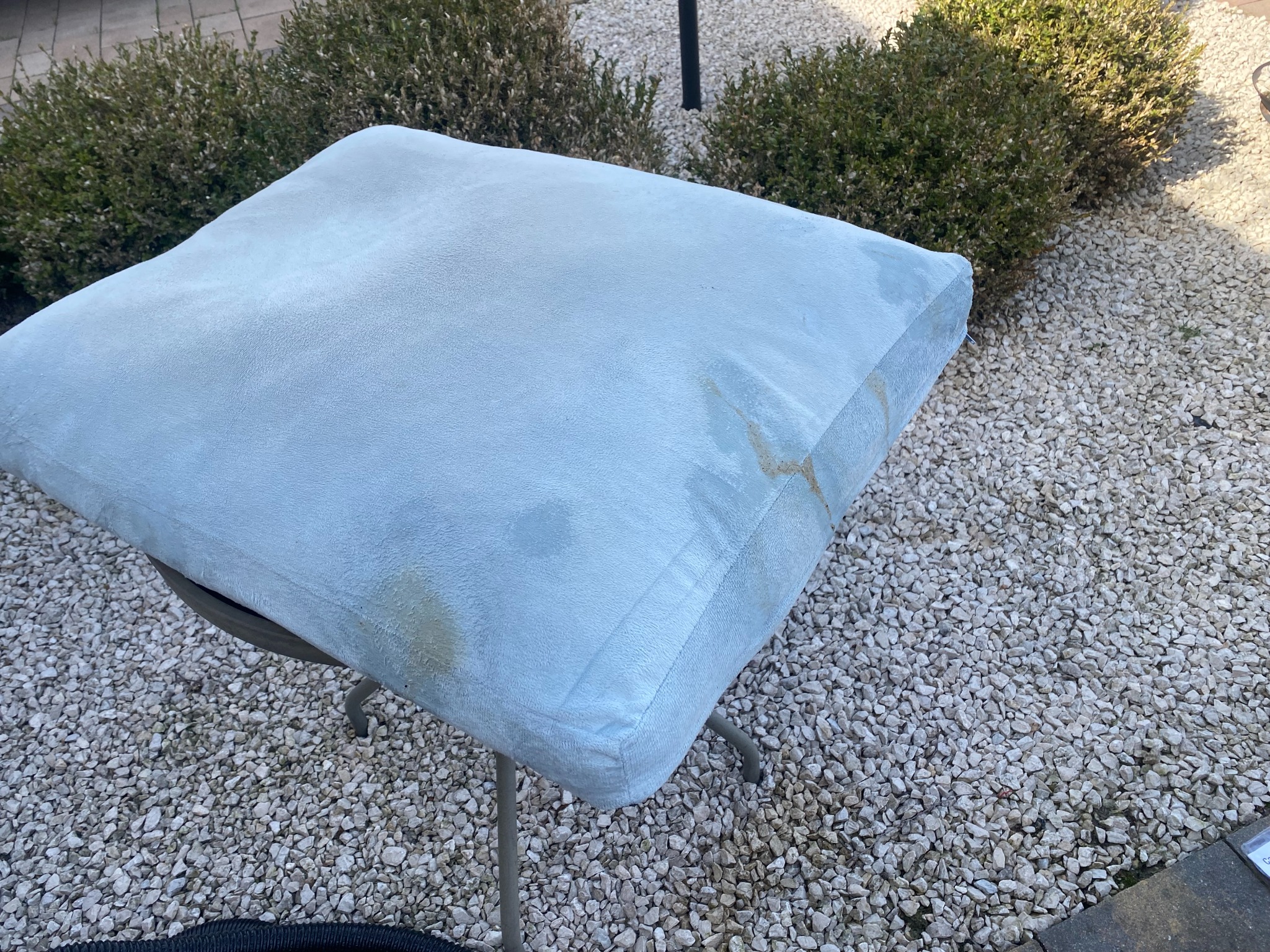 Jasnoniebieska poduszka na krześle ogrodowym z widocznymi plamami, ustawiona na żwirowym podłożu w otoczeniu zieleni.