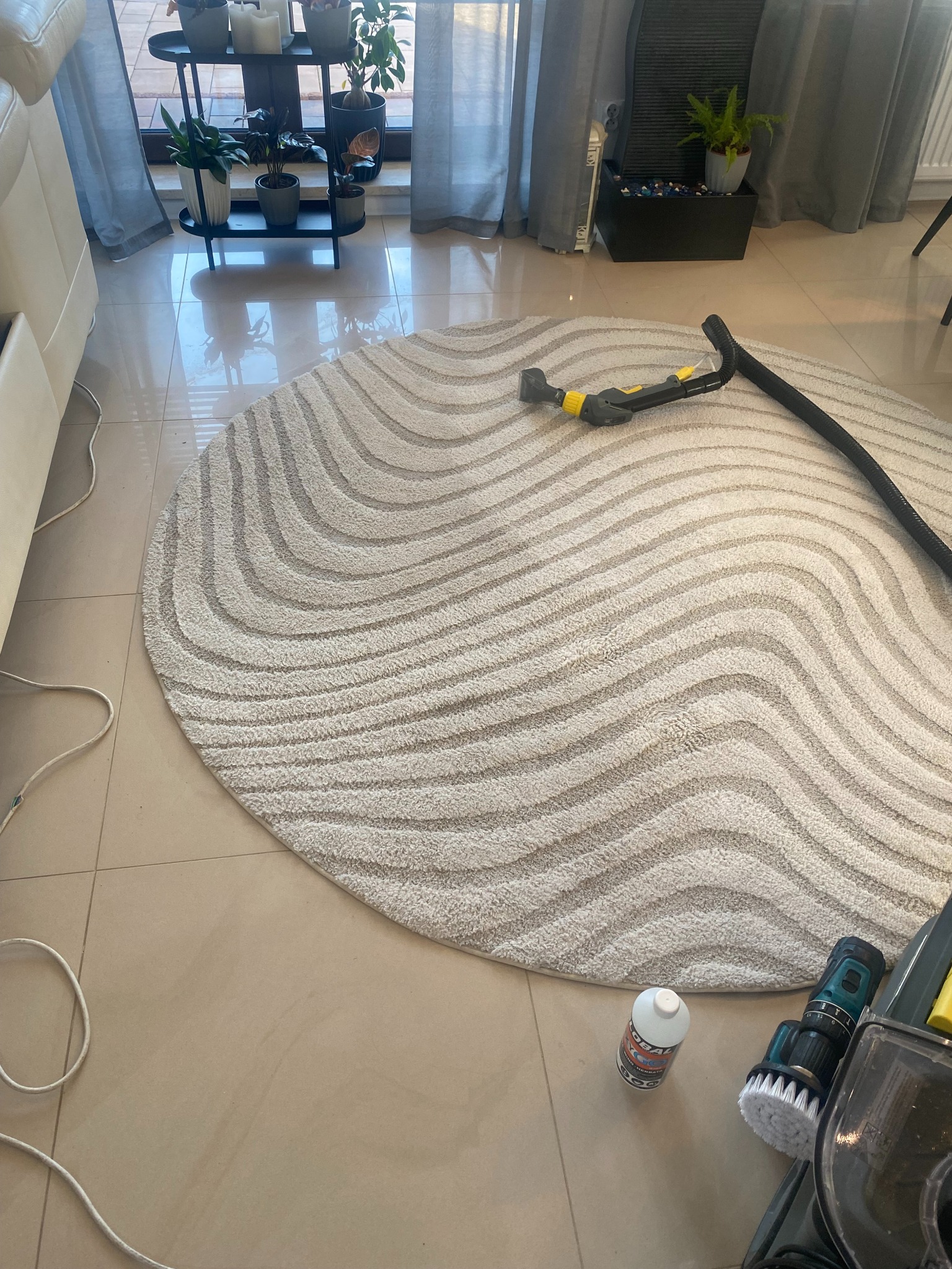 Okrągły dywan w fale czyszczony odkurzaczem piorącym. Na podłodze butelka płynu i szczotka do czyszczenia. Widok z góry, oświetlenie dzienne.