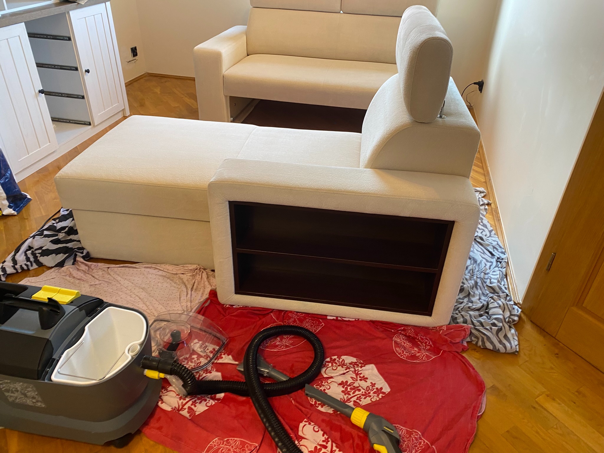 Jasna sofa modułowa częściowo przykryta tkaniną, obok odkurzacz piorący; widoczne elementy wnętrza, drewniana podłoga i biała szafka.